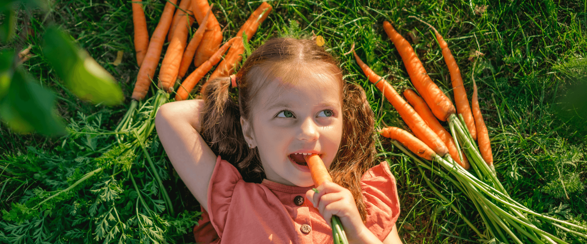 Petite fille allongée dans l'herbe, entourée de légumes, croquant une carotte - Header