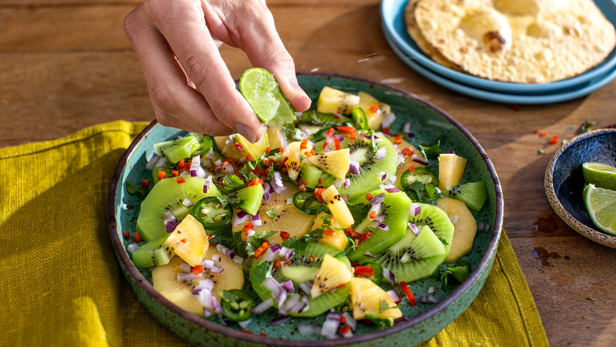 Hand squeezing lime juice over salad of green and golden kiwi on wooden table